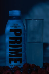 foto de botella prime con fondo azul, un baso de tubo y frutos rojos